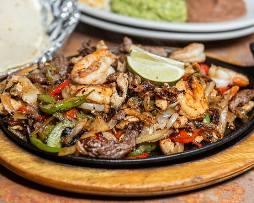 Sizzling fajitas with shrimp, beef, chicken, peppers, onions, and a lime wedge on a hot platter.