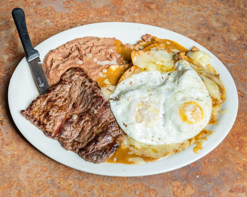 Grilled steak, refried beans, and two fried eggs over cheesy enchiladas with a steak knife.