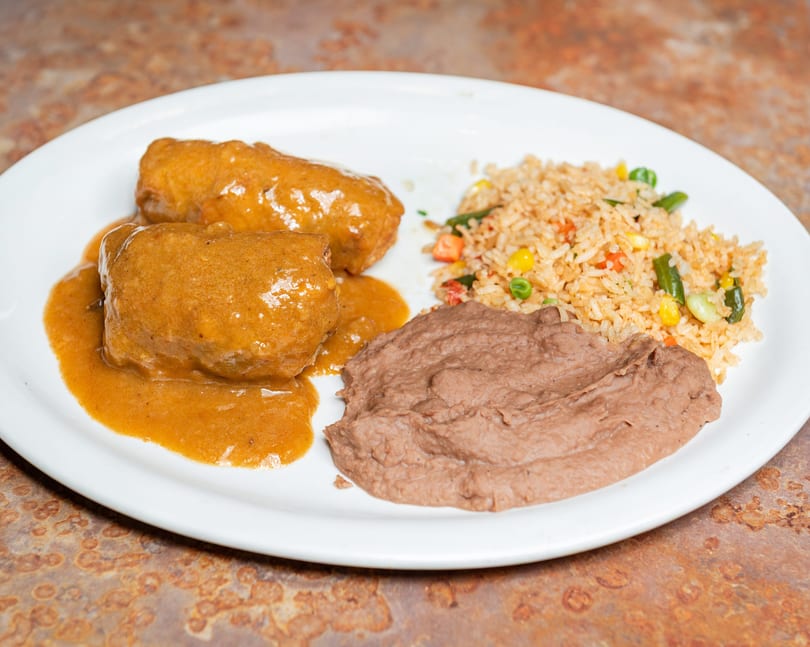 Two items in brown sauce, rice with vegetables, and refried beans on a white plate.