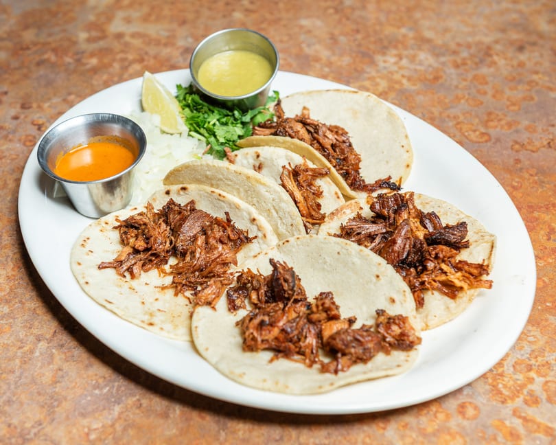Six shredded meat tacos with two dipping sauces, chopped onions, cilantro, and a lime wedge.