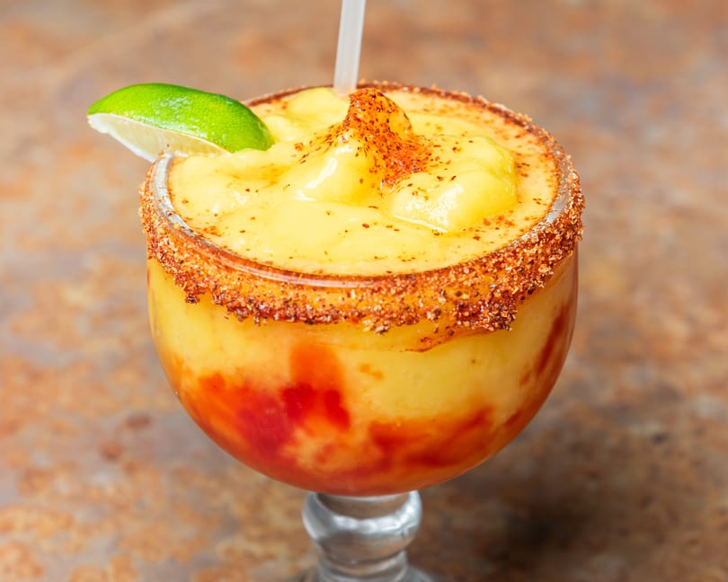 Frozen mango drink with red swirls, chili-lime rim, and a fresh lime wedge.