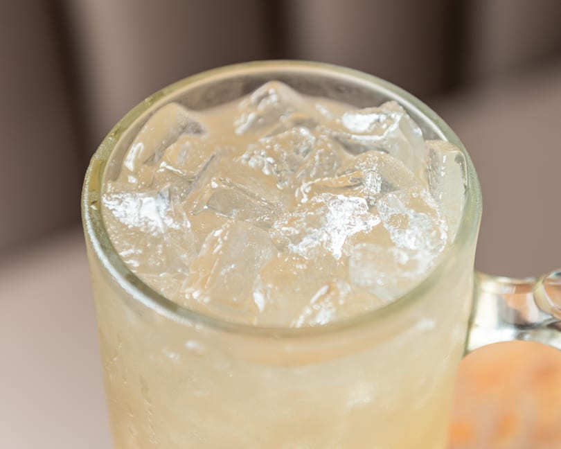 Glass mug filled with ice cubes and a light yellow drink, showing condensation.