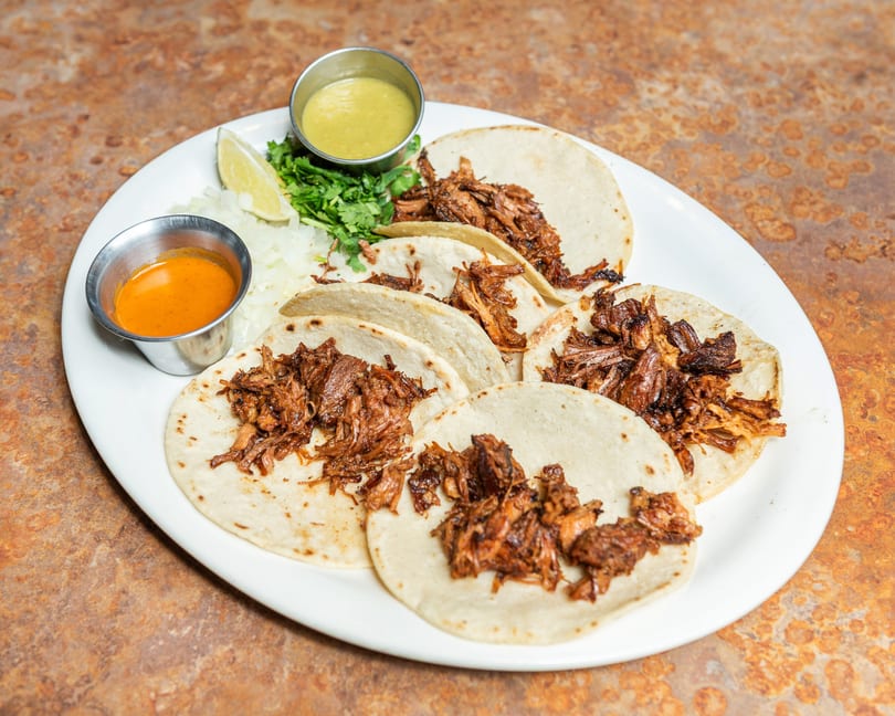Plate of six shredded meat tacos with two sauces, lime, cilantro, and diced onions.