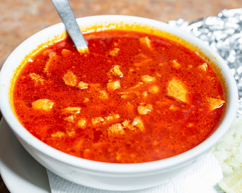 Vibrant red menudo soup with meat chunks and a spoon, served with onions and lime.